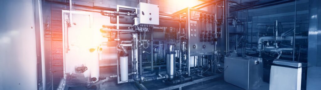 L’image montre une salle technique industrielle dédiée au traitement de l’eau, équipée de multiples dispositifs et tuyauteries. Au centre de la scène, un système de filtration complexe est monté sur une structure métallique robuste. Ce système comprend plusieurs filtres verticaux sous pression, des pompes motorisées, des manomètres, et des vannes de régulation montées en série sur des conduites horizontales et verticales en PVC ou en inox. À droite, on distingue un panneau de contrôle avec des cadrans analogiques, des tuyaux transparents probablement destinés au contrôle visuel des débits ou des niveaux, et une boîte électrique de commande montée sur le châssis métallique. Des câbles et tuyaux électriques sont également visibles, suspendus ou fixés à la structure. L’éclairage bleuâtre donne à la pièce une ambiance stérile et technique, avec un effet de halo lumineux chaud au centre gauche, simulant un éclairage LED intense ou un effet visuel volontairement accentué. Le sol est recouvert d’un revêtement lisse, adapté à un environnement industriel ou agroalimentaire. À l’arrière-plan, on distingue des parois métalliques réfléchissantes et d’autres éléments de tuyauterie, suggérant que cette salle fait partie intégrante d’une installation plus vaste. Sur la gauche de l’image, un espace blanc plus flou semble être l’entrée ou un couloir adjacent à la zone technique.